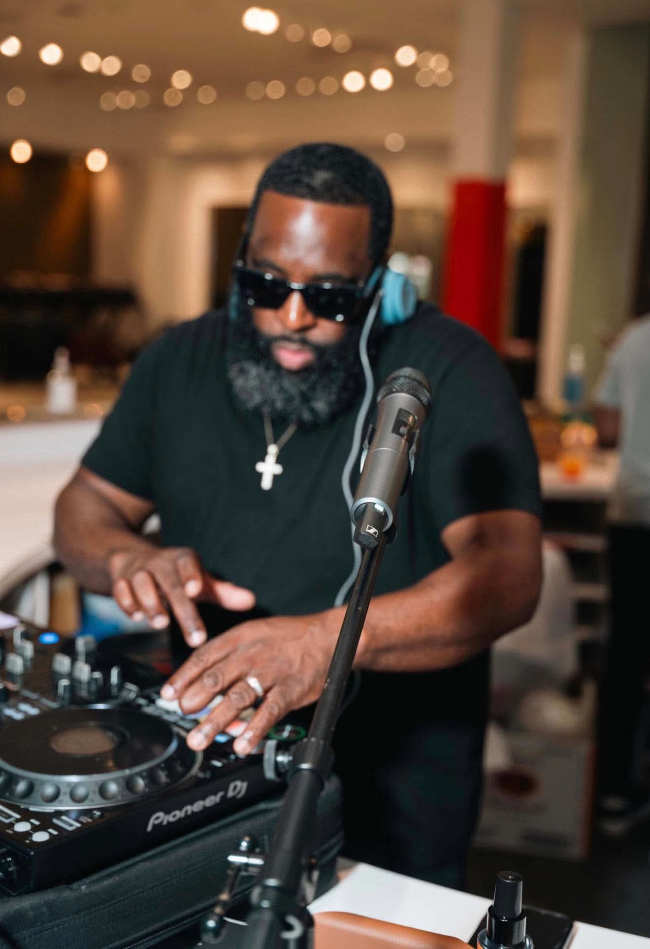 Stevie Pugh mixing on Pioneer DJ decks with headphones and sunglasses, a microphone on a stand in front of him
