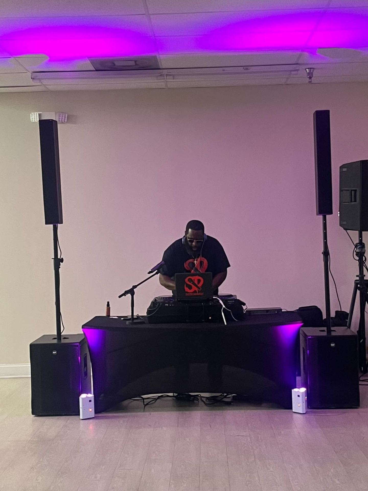 A full DJ production in an event hall: DJ Stevie Steve behind the booth, flanked by two column speakers on subwoofers with purple uplighting